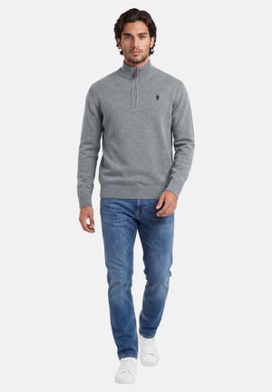 Man walking wearing a grey quarter-zip sweater, blue jeans, and white sneakers against a plain white background.