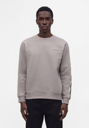 Jeune homme portant un sweat-shirt gris clair avec un logo sur la poitrine et un ruban rayé sur les manches, debout devant un fond blanc.
