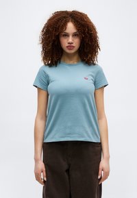 Νεαρή γυναίκα με σγουρά μαλλιά, που φοράει γαλάζια κοντομάνικη μπλούζα και σκούρο καφέ παντελόνι,  στέκεται μπροστά σε απλό φόντο.