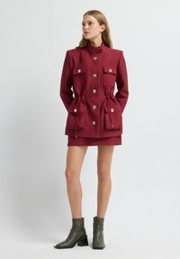 Donna che indossa una giacca rossa texturizzata con grandi bottoni e tasche, abbinata a una gonna coordinata e stivali alla caviglia verde oliva.