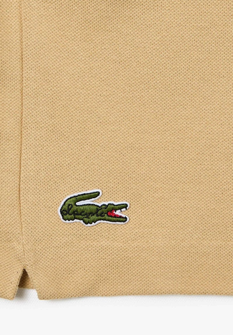 Polo de punto beige con tejido texturizado, que presenta un logo de cocodrilo bordado en verde con acentos rojos en el pecho.