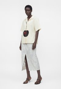 Femme portant une chemise crème à manches courtes avec un grand motif cerise et une jupe midi en dentelle florale blanche avec une fente à l'avant.