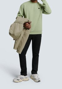 Ljusgrön sweatshirt med ribbade muddar, beige jacka över armen, svarta byxor och kraftiga vita sneakers med grå accenter.