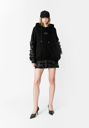 Sudadera negra oversized con detalles de cinta texturizada en las mangas, combinada con una falda negra a juego. La modelo también lleva una gorra negra y zapatillas.