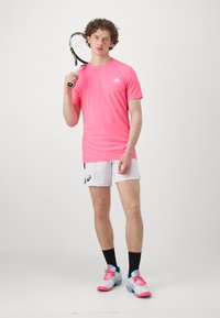 Pink athletic t-shirt, white shorts with logo, black socks, and multicolored tennis shoes. Holding a tennis racket.