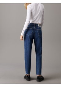 Kvinna som bär högmidjade mörkblå jeans och en vit långärmad crop top, som står mot en enfärgad grå bakgrund, vänd bort.