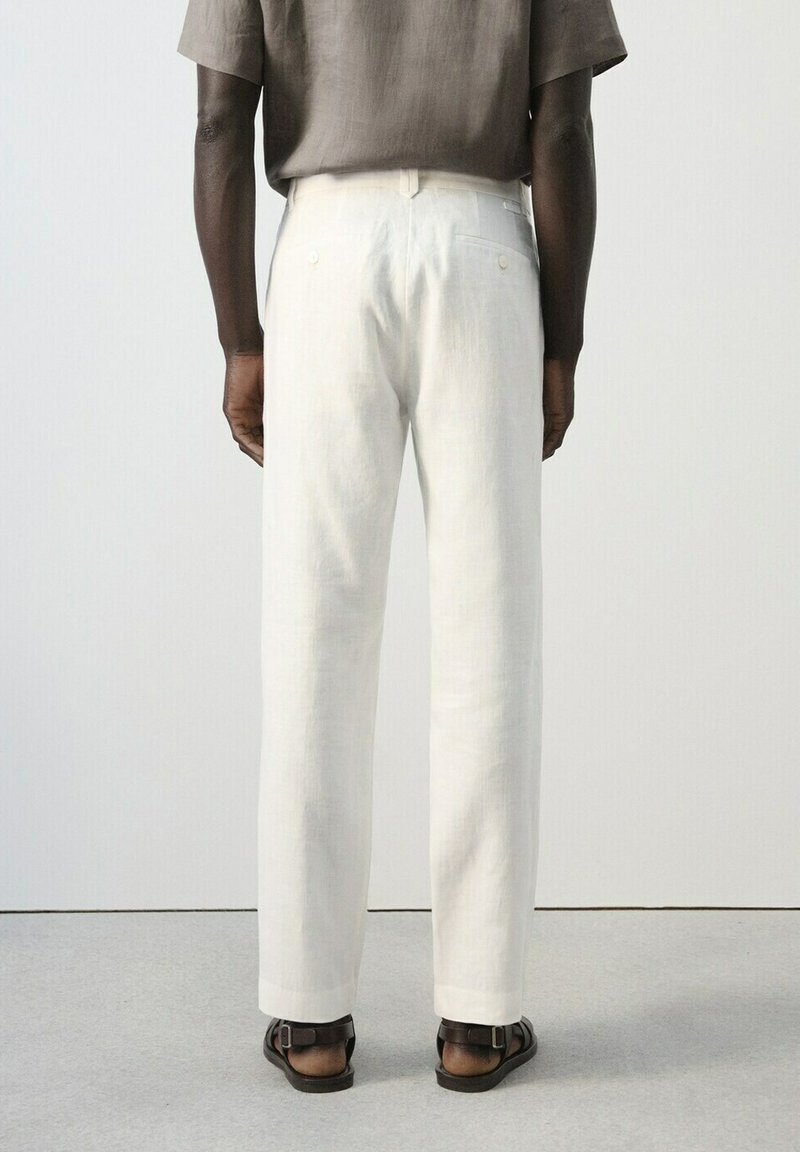 Homme portant un pantalon blanc à jambes droites, des sandales foncées et une chemise ample à manches courtes marron, debout de dos sur un sol gris.