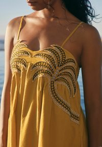 Robe jaune avec des bretelles fines, ornée de motifs de feuilles de palmier brodés en noir et blanc sur le corsage. Tissu doux avec un design fluide.
