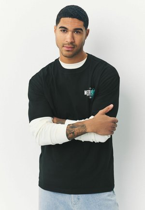 Sort kortærmet t-shirt med rund halskant og logo detaljer i turkis, lagdelt over en hvid langærmet skjorte; blå denim bukser.
