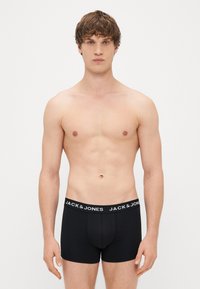 Črne bokserice s tesnim fitom in elastičnim pasom, ki nosi belo blagovno znamko. Tkanina se zdi gladka in raztegljiva.