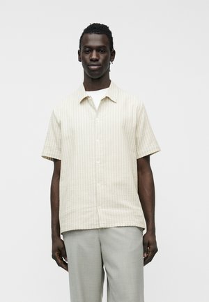 Άνδρας φοράει μπεζ πουκάμισο με κοντά μανίκια και κουμπιά πάνω από λευκό μπλουζάκι και ανοιχτό γκρι παντελόνι, στέκεται μπροστά σε απλό λευκό φόντο.