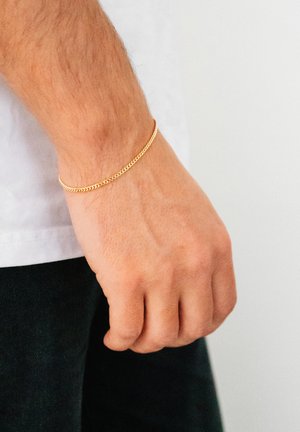 Gold chain bracelet resting on a wrist, featuring a woven design and a smooth texture, contrasting against the skin and clothing.