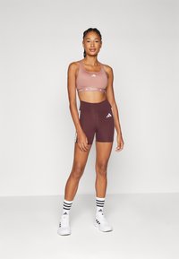 Sport-bh i lys rosa med hvit logo og bånd, kombinert med dyp burgunder shorts med hvite striper og logoer. Hvite joggesko og stripete sokker.