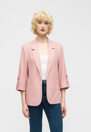 Femme blonde portant un blazer rose clair aux manches retroussées trois quarts, un haut blanc et un jean bleu sur un fond blanc uni.