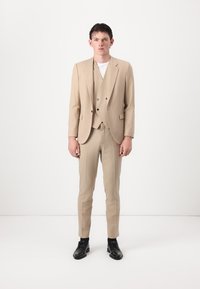 Completo tre pezzi beige su misura con rever a punta, pantaloni slim fit e bottoni. Indossato con una camicia bianca e scarpe nere.