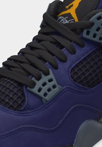 Κοντινό πλάνο ενός μωβ και μαύρου αθλητικού παπουτσιού με μαύρα κορδόνια, γκρι οπές κορδονιών, πάνελ από δίχτυ και κίτρινο λογότυπο Jumpman στη μαύρη γλώσσα.