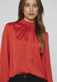 Sarkans garās piedurknes blouse ar smocked apkakli, izgatavota no gludas, spīdīgas auduma. Modelis to valkā iešūtu melnās biksēs ar pogu detaļu.