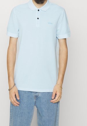 Mies, joka pukeutunut vaaleansiniseen lyhythihaiseen poolopaitaan, jossa on kaksi mustaa nappia, ja vaaleansinisiin farkkuihin, seisoo rentoina käsivarsina sivuillaan.