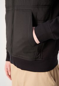 Personne portant une veste noire zippée avec une main dans la poche avant, associée à un pantalon beige.