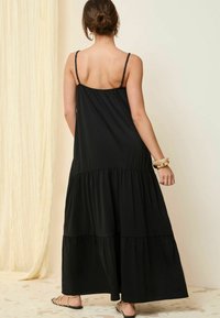 Svart maxiklänning med spaghetti-remmar, med ett våningsdesign och plisserad textur. Bärs med platta sandaler och ett armband.