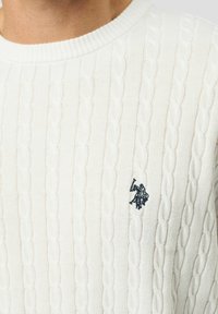 Prim-plan al unei persoane care poartă un pulover alb cu model de cablu tricotat și un mic logo negru brodat pe piept.