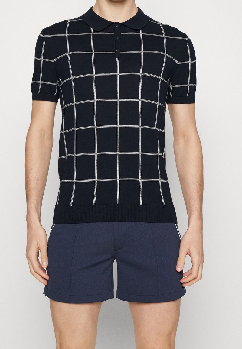 Homme portant un polo noir à manches courtes avec un motif quadrillé blanc et un short bleu marine foncé, debout devant un fond uni.