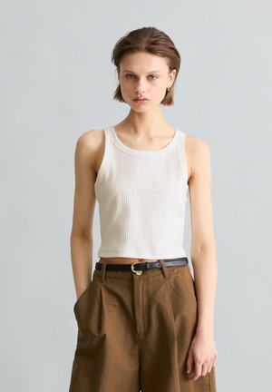 Jeune femme portant un haut blanc sans manches côtelé et un pantalon marron taille haute avec une ceinture noire, debout devant un fond gris uni.