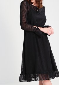 Černé šaty s tečkovanou texturou, dlouhými rukávy a volánkovým lemem. Mají zavazování kolem krku a na pase s řasením pro splývavý siluetu.