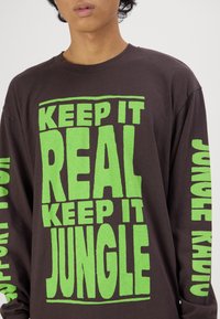 Μακρυμάνικο καφέ βαμβακερό μπλουζάκι με έντονα πράσινα γράμματα που αναγράφουν "ΔΙΑΤΗΡΗΣΕ ΤΗΝ ΠΡΑΓΜΑΤΙΚΟΤΗΤΑ, ΔΙΑΤΗΡΗΣΕ ΤΗ ΖΟΥΓΚΛΑ" στο στήθος και τα μανίκια.