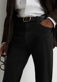 Persoon met zwarte broek, wit overhemd en donkere jas met hand in zak, houdt een bril in de andere hand.
