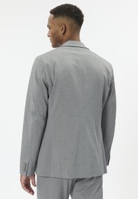 Vyras su trumpais garbanotais plaukais, apsirengęs pritaikytu pilku kostiumu, pavaizduotas iš nugaros ant paprasto balto fono.