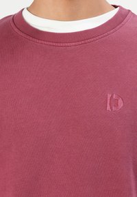 Detailný záber na osobu, ktorá má na sebe vínový sveter s kruhovým výstrihom, na ktorom je na ľavej hrudi malé vyšívané logo, cez biele spodné tričko.