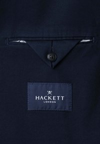 Marineblauer Stoff mit einer geknöpften Klappentasche und einem marineblauen Label mit der Aufschrift "Hackett London" und einem Logo mit gekreuzten Schlägern darüber.