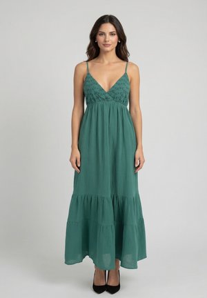 Mujer con vestido largo verde sin mangas con bordado floral en el corpiño y tacones altos negros, de pie contra un fondo liso y claro.