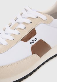 Hvit sneakers med en beige semsket tå og brune lærdetaljer. Inkluderer hvite snørebånd og en teksturert sort gummisåle. BOSS-logoen vises.