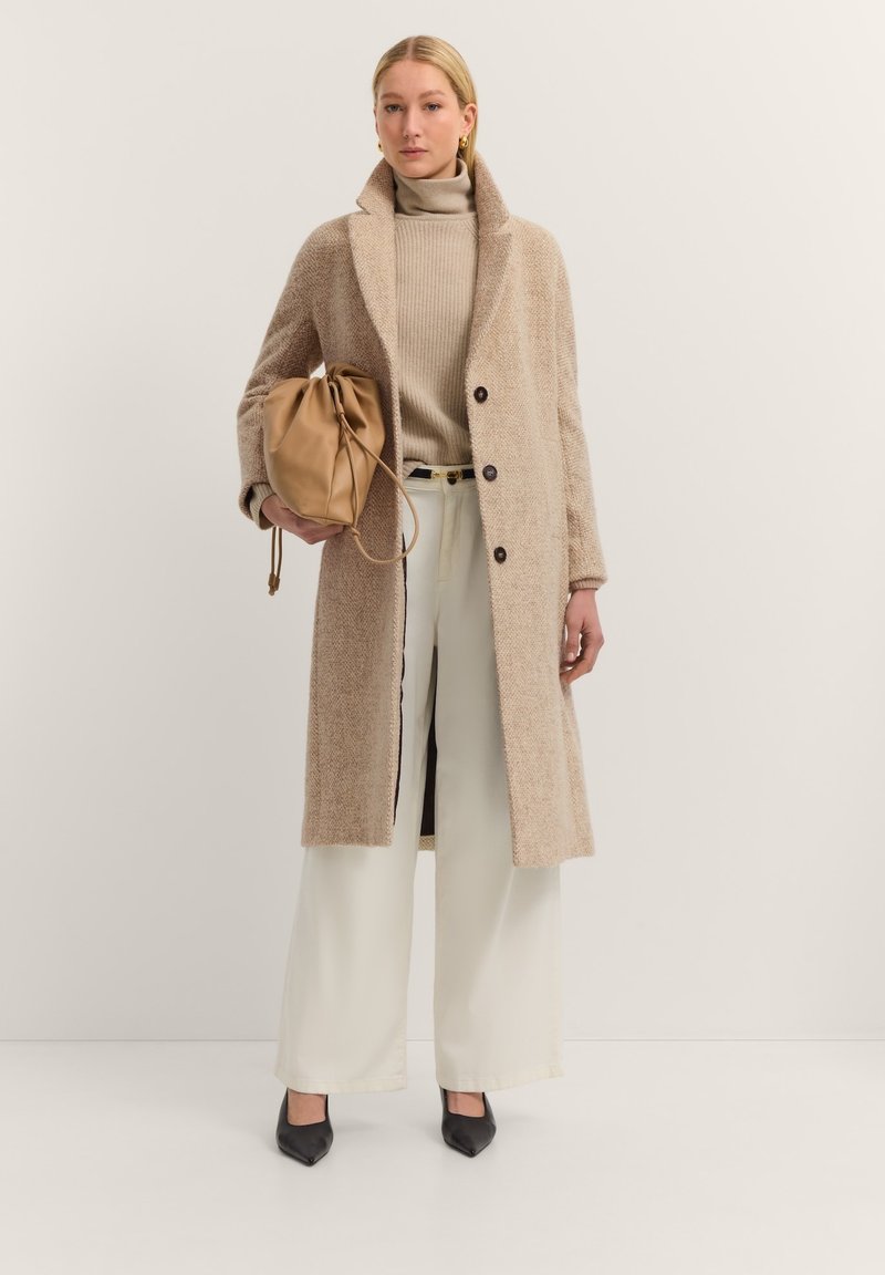 Beige langer Wolle-Mix-Mantel mit Kragen, zwei Knöpfen und strukturiertem Stoff, kombiniert mit einer beige Tasche, einem cremefarbenen Rollkragenpullover und weiten Hosen.