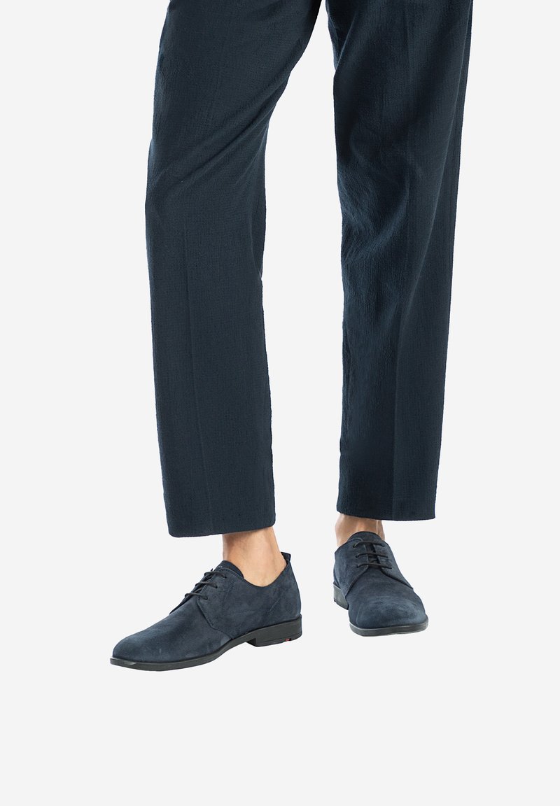Chaussures en daim bleu marine avec une texture lisse, à lacets, et un petit talon, portées avec un pantalon sombre ample.