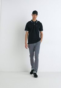 Μαύρο πόλο μπλούζα με λευκό γιακά και φινίρισμα, γκρι παντελόνι και μαύρα αθλητικά παπούτσια. Το μοντέλο φοράει μαύρο καπέλο και στέκεται σε λευκό φόντο.