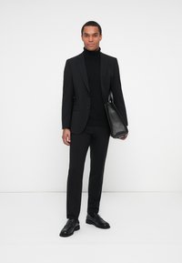 Completo nero con design aderente, abbinato a un dolcevita nero. L'individuo tiene una borsa in pelle nera, completata da scarpe nere.