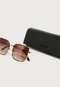 Sluneční brýle Calvin Klein s obroučkami ve vzoru želvoviny a hnědými gradientními čočkami, vedle nich černé ochranné pouzdro s logem.