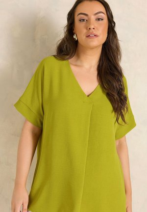 Kobieta o długich brązowych włosach, ubrana w limonkową bluzkę z krótkim rękawem i dekoltem w serek oraz złote kolczyki-koła, na neutralnym tle.