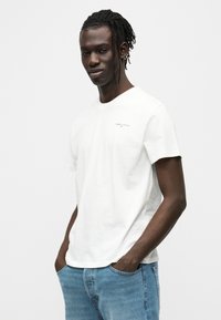 Άσπρο βαμβακερό T-shirt με στρογγυλή λαιμόκοψη και κοντά μανίκια. Μικρό γκρι λογότυπο στο αριστερό στήθος. Συνδυάζεται με ανοιχτό μπλε τζιν παντελόνι.