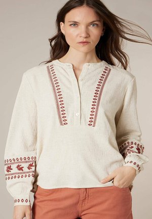 Femme aux cheveux bruns portant un chemisier crème texturé avec une broderie rouge et un pantalon couleur rouille, debout avec une main dans la poche.