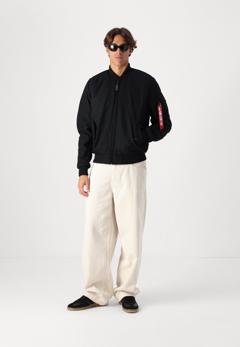 Schwarze Bomberjacke mit Reißverschluss vorne und gerippten Bündchen, mit einem roten Etikett am linken Arm. Kombiniert mit cremefarbenen weiten Hosen und schwarzen Sneakers.