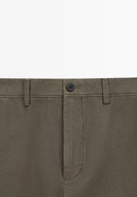Gros plan sur la ceinture d'un pantalon vert olive avec un seul bouton noir, des passants pour ceinture et une braguette zippée recouverte.