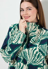 Långärmad topp med hög krage, med ett botaniskt mönster i grönt och marinblått på en krämfärgad bakgrund, tillverkad av mjukt material.