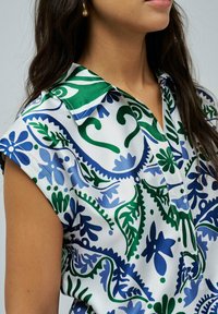 Blusa de manga corta en una tela similar al satén, con un patrón floral en azul y verde sobre un fondo blanco y un cuello tipo camisero.