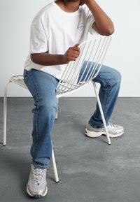 Valkoinen puuvillainen T-paita, pienellä sydänlogolla, siniset farkut ja harmaat tennarit hopeisilla yksityiskohdilla, istuen valkoisella metallisella tuolilla.