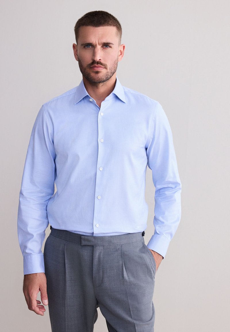 Homme aux cheveux courts et à la barbe, portant une chemise bleu clair boutonnée et un pantalon gris, debout devant un fond uni.