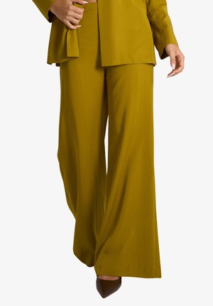 Persona che indossa pantaloni giallo senape a gamba larga con blazer abbinato e scarpe scure a punta, mano che sistema il blazer.